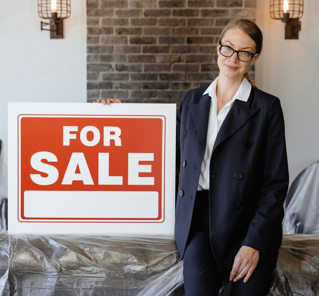Businesswoman in suit with glasses holding a 'For Sale' sign indoors against a brick wall.