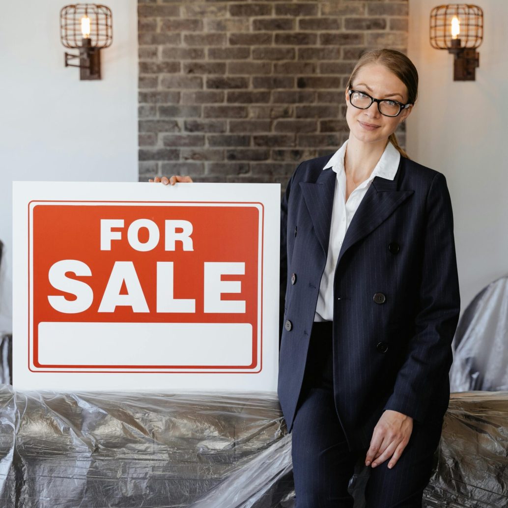 Businesswoman in suit with glasses holding a 'For Sale' sign indoors against a brick wall.