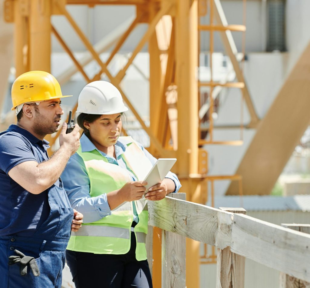 Two construction workers in hardhats discuss plans using a tablet and walkie talkie on-site.