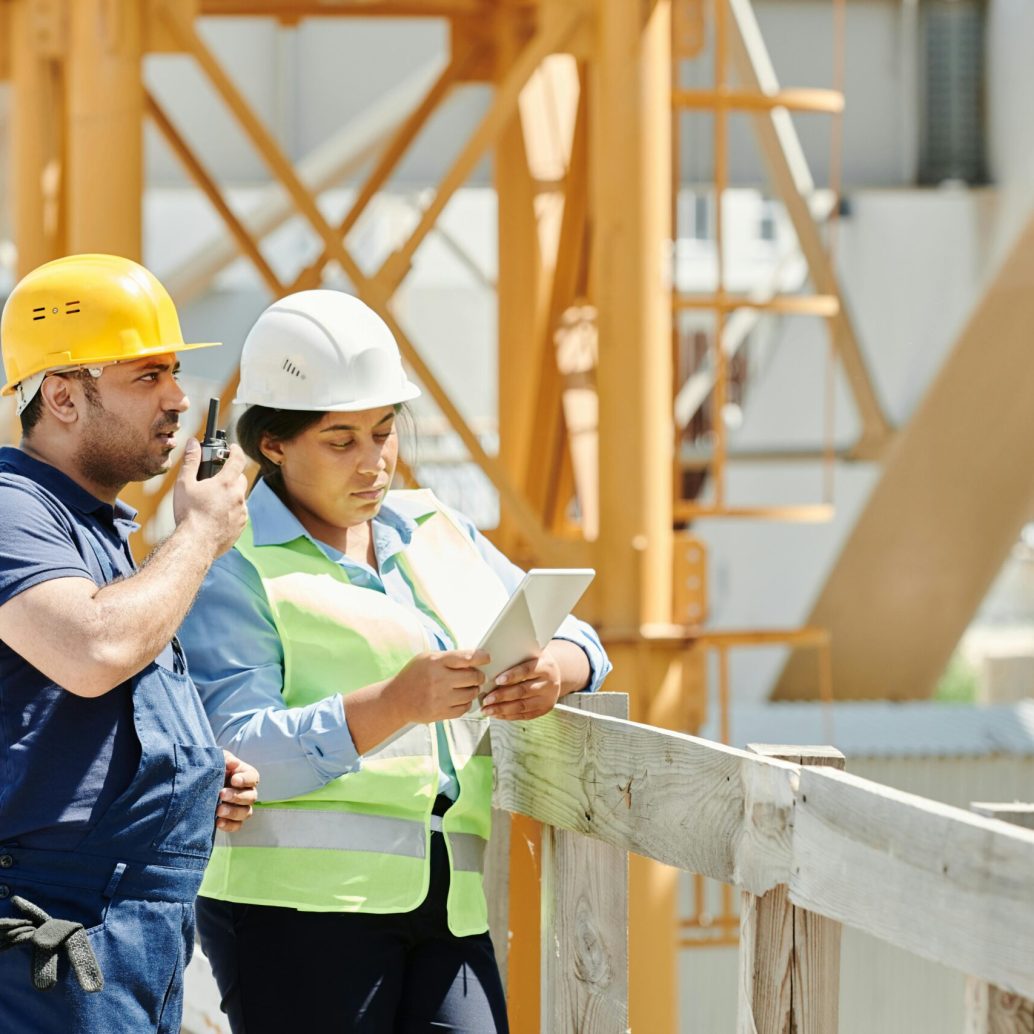 Two construction workers in hardhats discuss plans using a tablet and walkie talkie on-site.